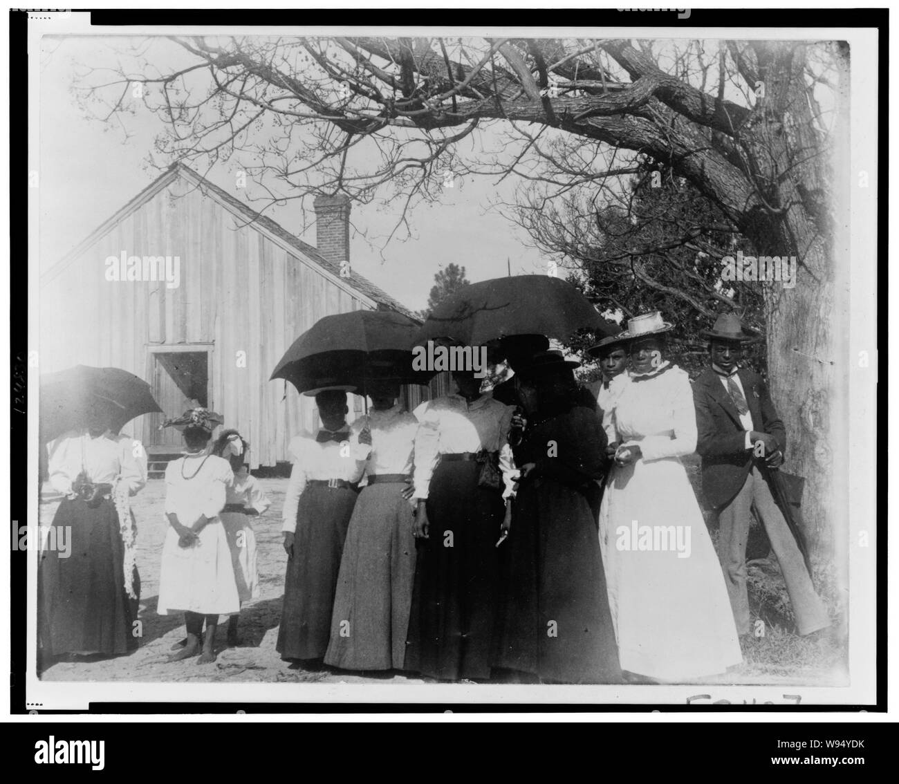 Afrikanische amerikanische Frauen, die Sonnenschirme Schatten von der Sonne zur Verfügung zu stellen, mit zwei Männern, und mit einem Gebäude (Kirche oder Haus der Begegnung) im Hintergrund Stockfoto Afrikanische amerikanische Frauen, die Sonnenschirme Schatten von der Sonne zur Verfügung zu stellen, mit zwei Männern, und mit einem Gebäude (Kirche oder Haus der Begegnung) im Hintergrund Stockfoto