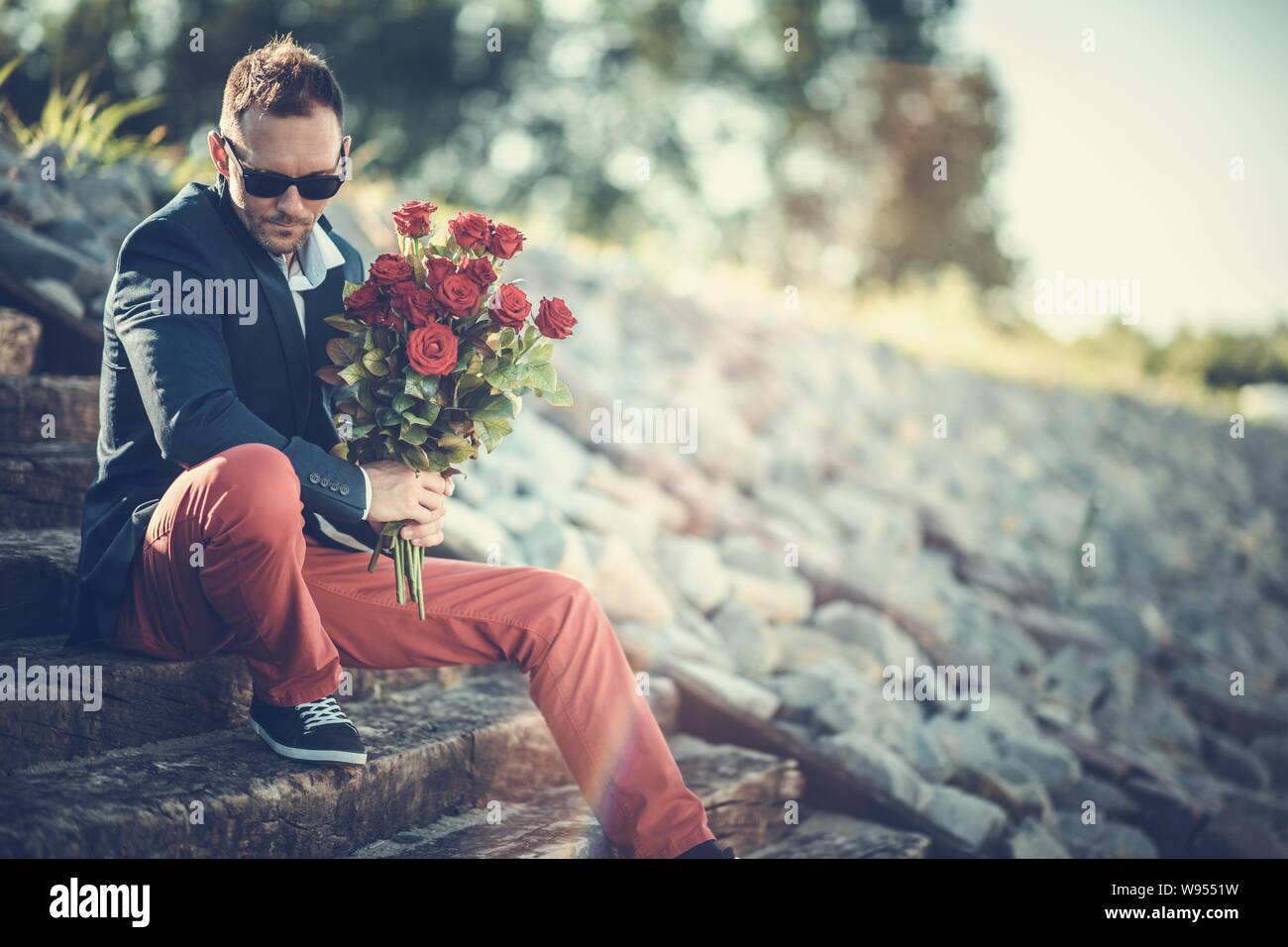 Kaukasischen Männern in seinem 30s warten auf Sein Date, während auf der Beton Treppe mit Blumenstrauß aus roten Rosen, frisch geschnitten. Stockfoto