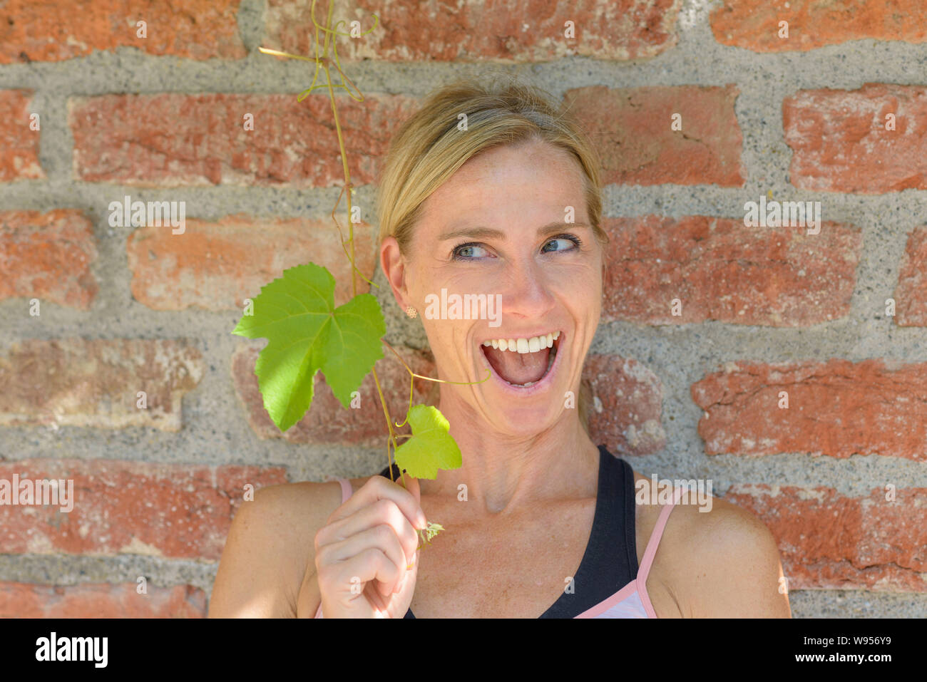 Verspielte Frau halten, frische grüne Blätter mit einem strahlenden Lächeln vor einem alten Äußeren Mauer Stockfoto