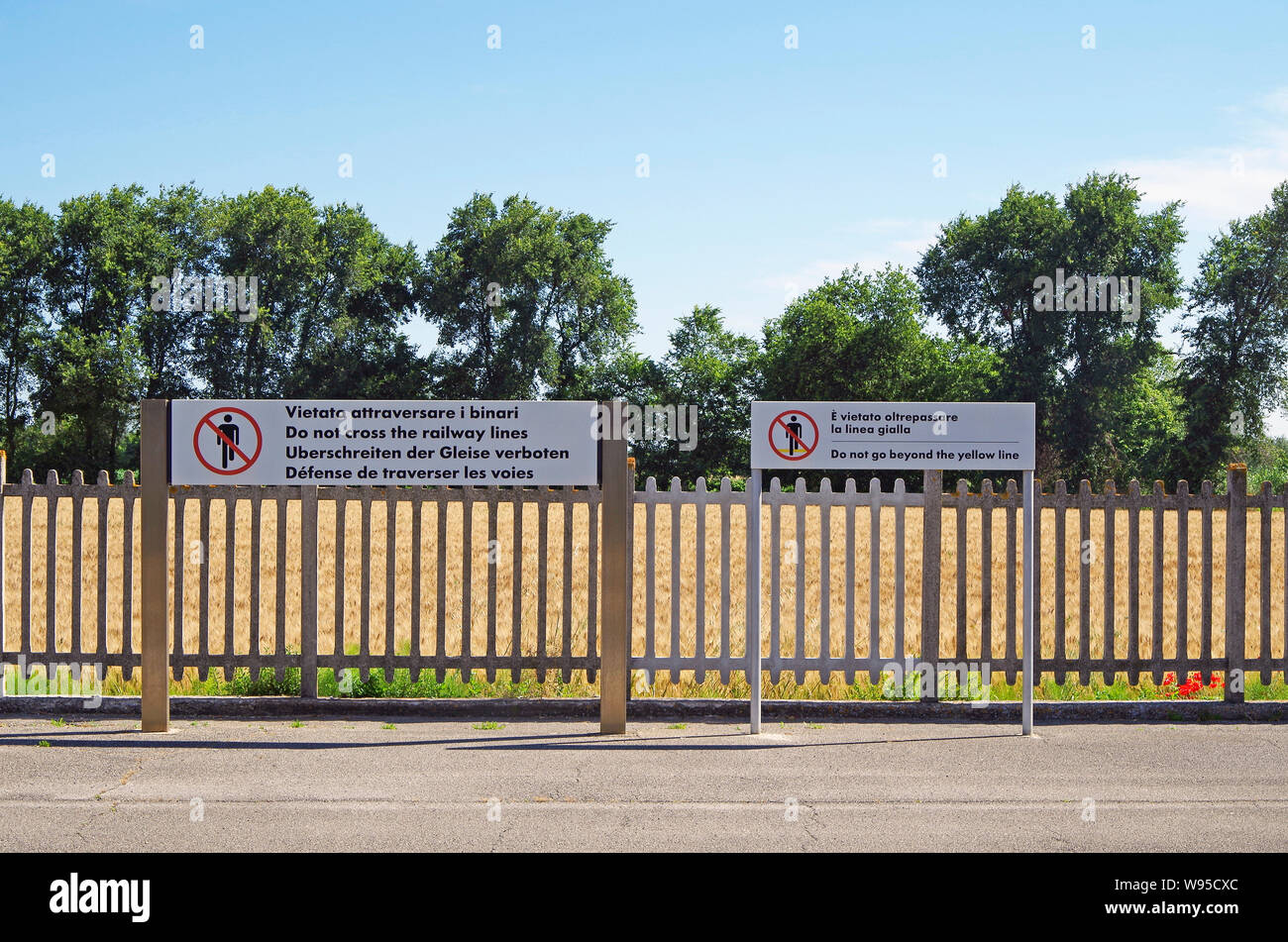 Zwei mehrsprachige Verbotsschilder dem Bahnübergang, der Gerste, die von hohen Bäumen, Lattenzaun, Loreto Station, Italienisch East Coast Line Stockfoto