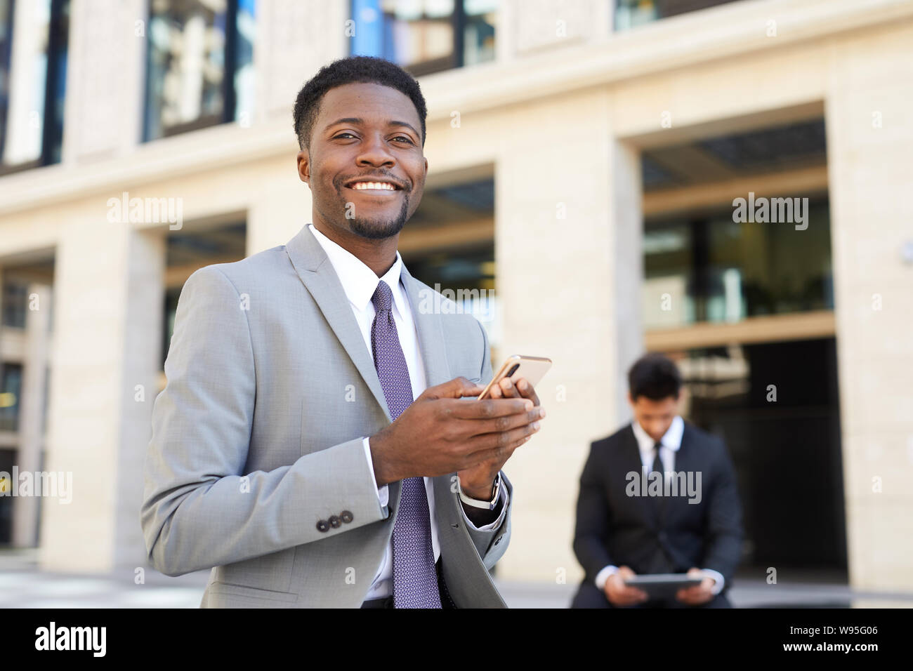 Portrait der Afrikanischen gut aussehender Geschäftsmann in Anzug mit Handy und lächelnd an der Kamera beim Stehen in der Stadt Stockfoto