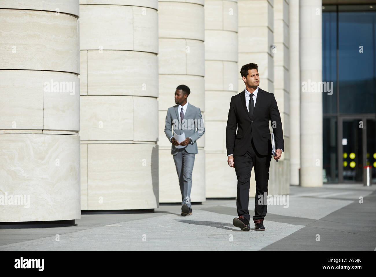 Ernsthafte zuversichtlich Geschäftsmann in schwarzen Anzug zu Fuß entlang der Straße in der Stadt mit anderen Mann im Hintergrund Stockfoto