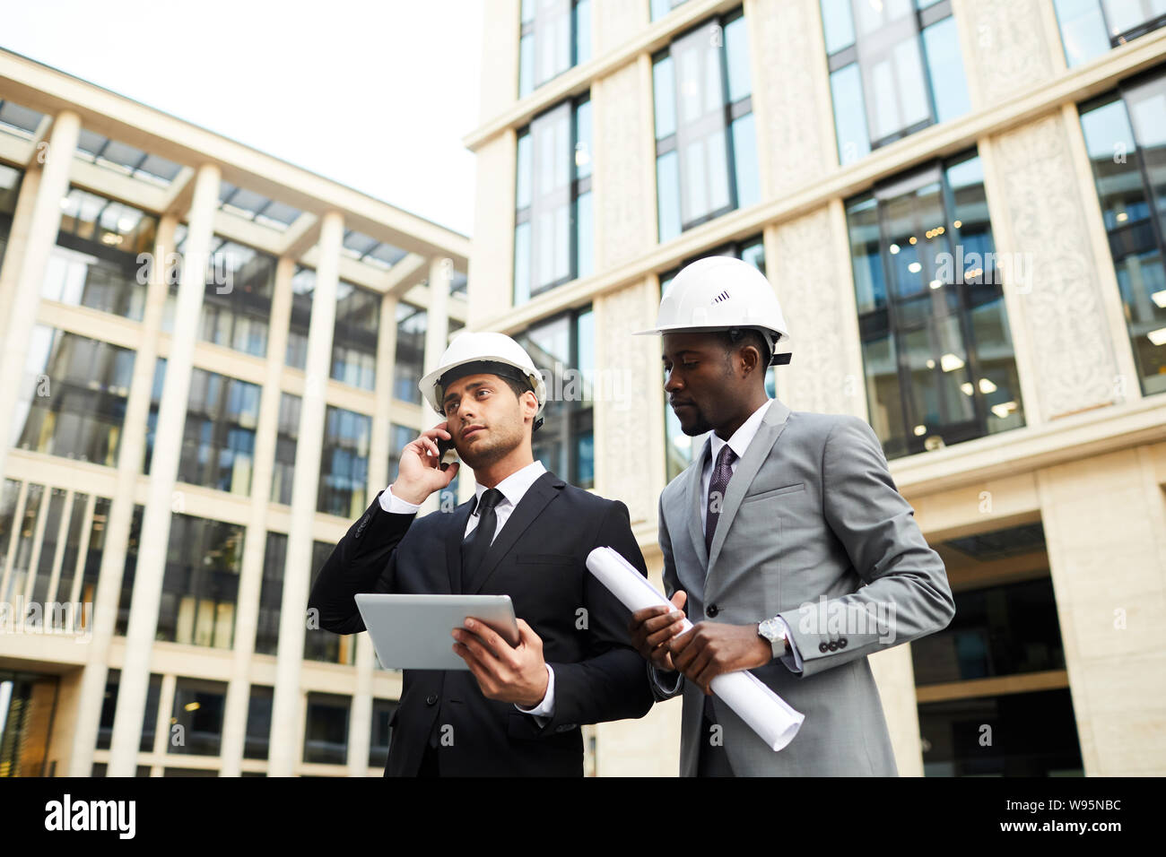 Junge Ingenieur in Anzug und Helm Gespräch am Handy mit seinem Kollegen in der Nähe von durch Sie in der Stadt arbeitet Stockfoto