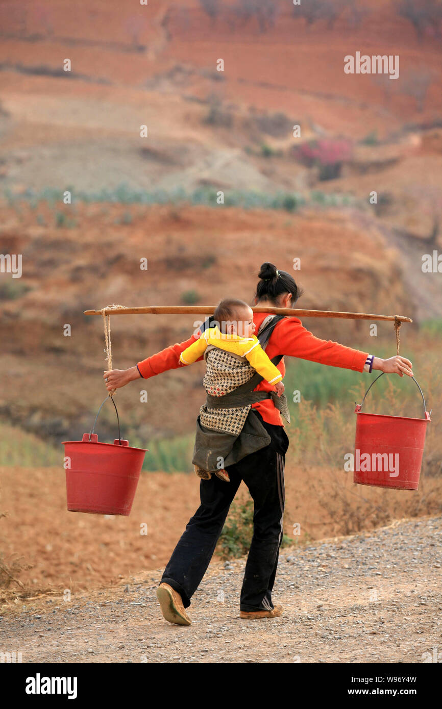 Eine chinesische Frau, die ihr Kind auf dem Rücken Schultern Eimer Wasser auf dem Weg zurück nach Hause von einer Wasserversorgung Station während einer Dürre in Yiliang Stockfoto