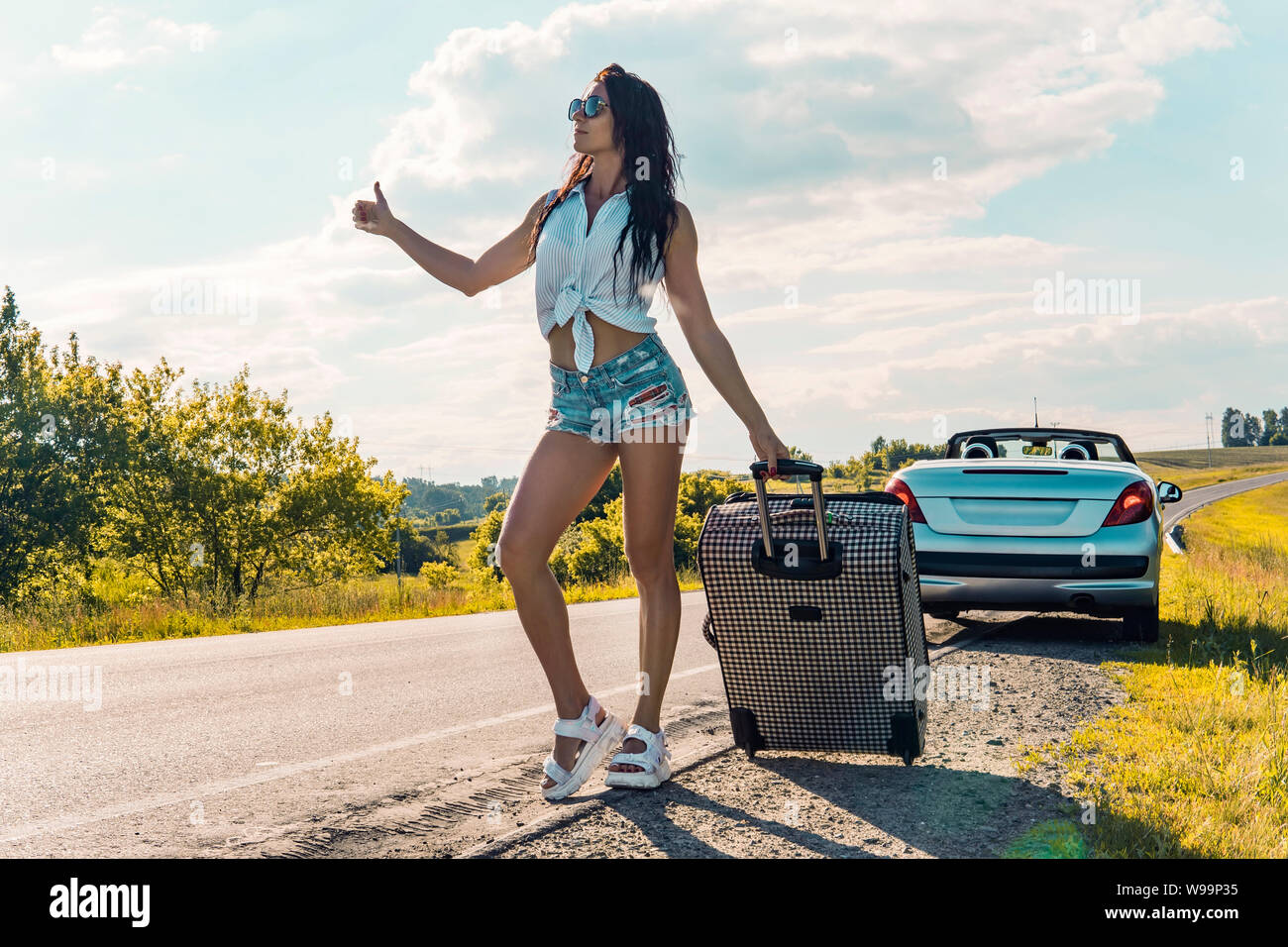 Frau mittleren Alters mit einer Reisetasche per Anhalter Fahren auf schmalen leere Landstraße, im Sommer auf dem Hintergrund einer kaputten Auto schöne Natur außerhalb der Stadt Stockfoto
