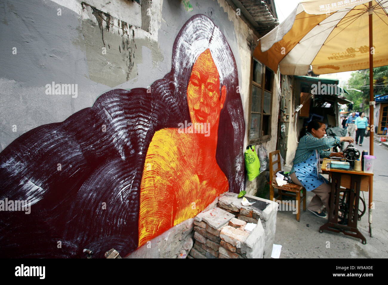 Ein chinesischer Schneider nutzt Ihre Nähmaschine neben einem Gemälde von einer Frau an der Wand eines Hauses in der Spur eines Slums in Shanghai, China, 26. Mai 2010. Stockfoto