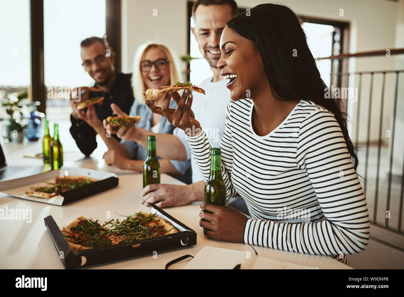 Lachende junge afrikanische amerikanische Geschäftsfrau, Sitzen mit Kollegen im Büro nach der Arbeit essen Pizza und trinken ein Bier Stockfoto