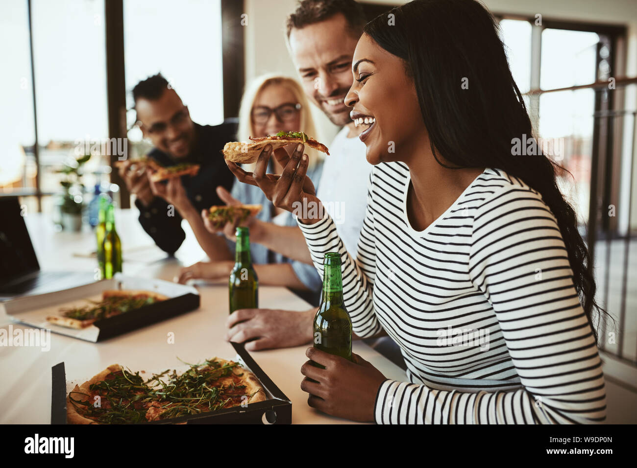 Lachende junge afrikanische amerikanische Geschäftsfrau sitzt in einem Büro mit Kollegen nach der Arbeit essen Pizza und trinken ein Bier Stockfoto