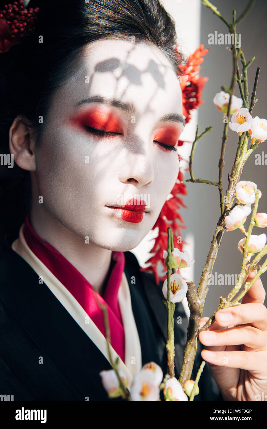 Wunderschöne Geisha mit roten und weißen Make-up und Sakura im Sonnenlicht Stockfoto