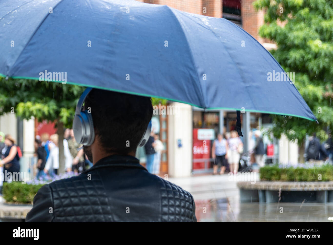 Ein Mann mit Kopfhörern, die Abdeckung vom Regen unter einem Sonnenschirm in einem Einkaufszentrum Stockfoto