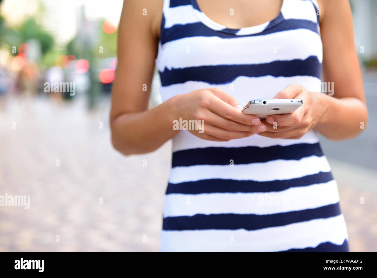 Mittelteil der jungen Frau SMS-Nachrichten auf dem Smartphone auf der Straße. Sie hält mobile Handy beim Gehen auf der Straße in der Stadt. Vorderansicht des Weibchens ist in gestreifter Kleidung. Stockfoto