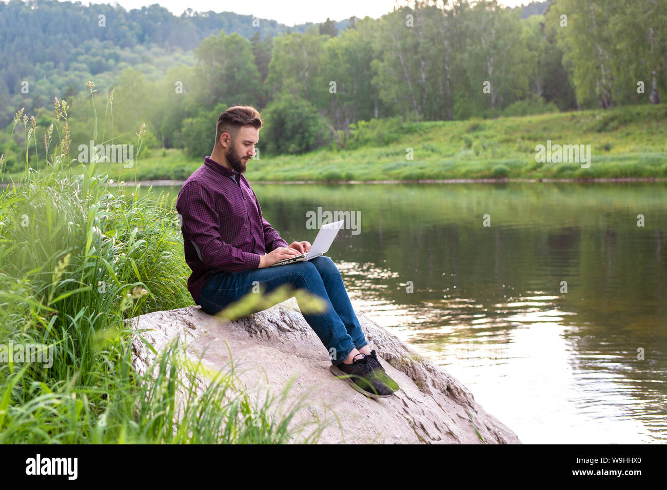 Bärtiger Mann, Freelancer sitzend mit einem Laptop am Fluss. remote arbeiten Konzept, freiberufliche Stockfoto