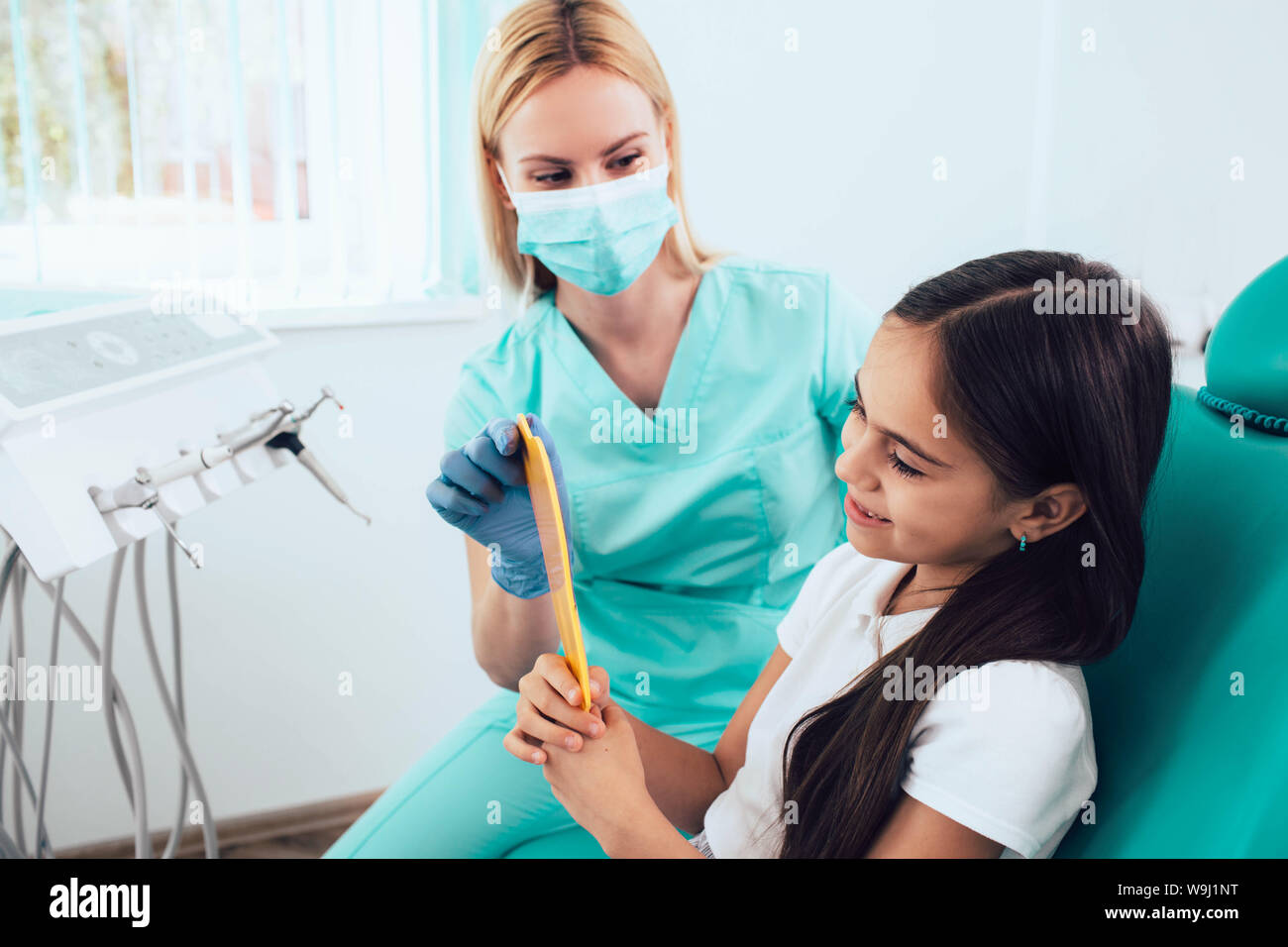 Kleines Mädchen mit Spiegel und lächelnd nach erfolgreicher Behandlung in der Zahnarztpraxis. Pädiatrische Zahnheilkunde Stockfoto