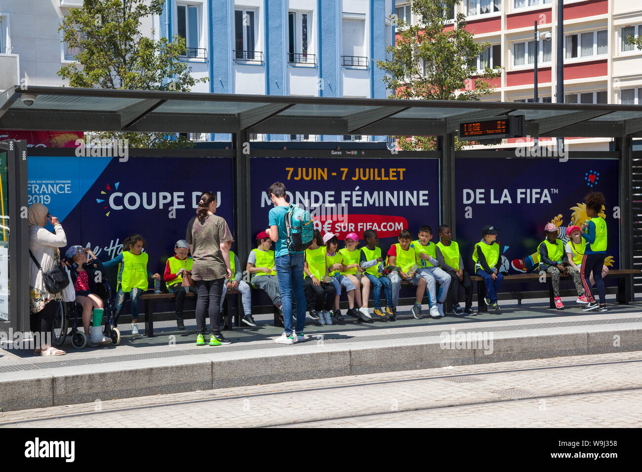 Schule Kinder in Gelb fluoreszierende Jacken warten Afür die Straßenbahn gegenüber dem Bahnhof in Le Havre, Frankreich mit einer Werbung für die Fifa Frauen Worl Stockfoto