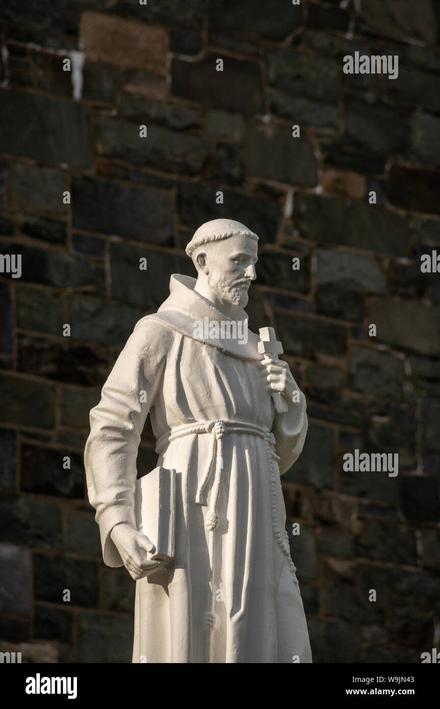 Religiöse Figur eines Heiligen Kreuz und Buch gegen Kirche stein Wand Fassade dunklen Hintergrund. Hl. Franz von Assisi Statue in Killarney, Irland. Stockfoto