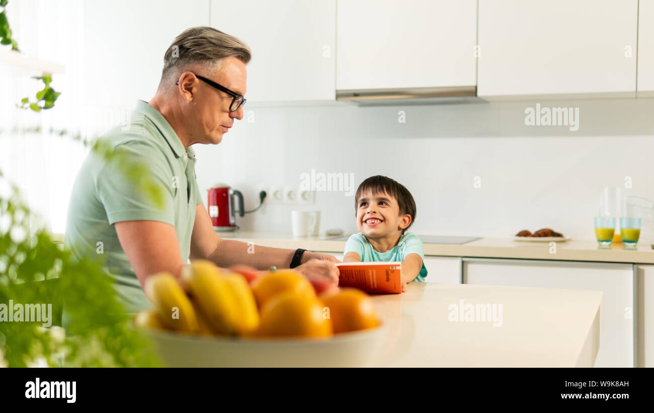 Der erwachsene Mann liest Buch zu seinem Sohn zu Hause, in der Küche. Happy Family im hellen Zimmer Stockfoto