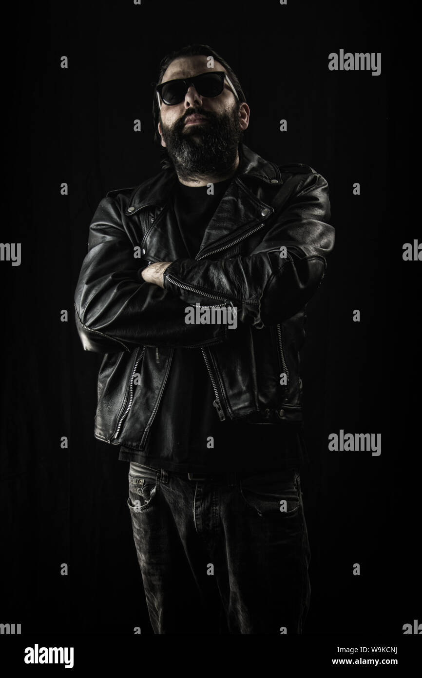 Harter Kerl in schwarzer Lederjacke und Sonnenbrille mit verschränkten Armen. Schuss gegen einen dunklen schwarzen Hintergrund. Stockfoto