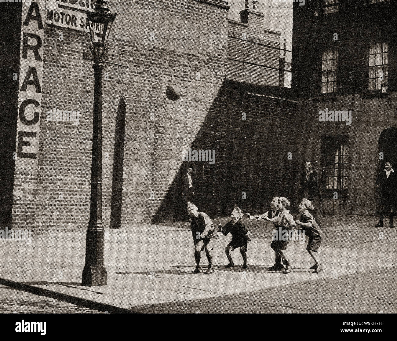 Circa 1940 - Britische Schule Kinder aus dem Norden Englands Fußball spielen in einem gepflasterten Platz neben der Straße, während die erwachsenen Nachbarn oder Väter auf. Stockfoto