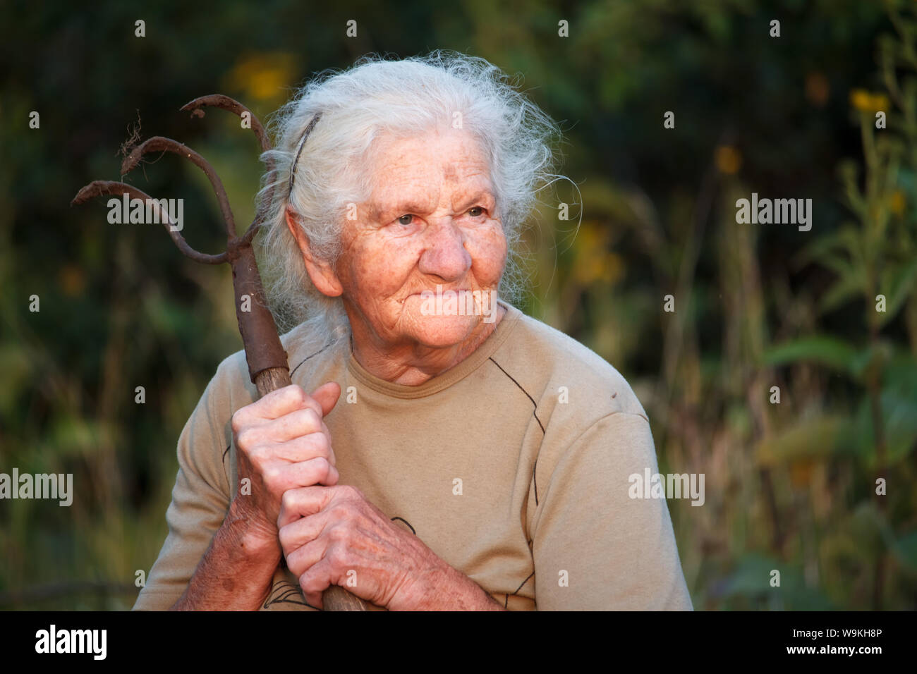 Closeup Portrait von einer alten Frau, die mit dem grauen Haar mit einem rostigen Mistgabel oder Chopper in ihre Hände, Gesicht in tiefe Falten, selektiver Fokus Stockfoto