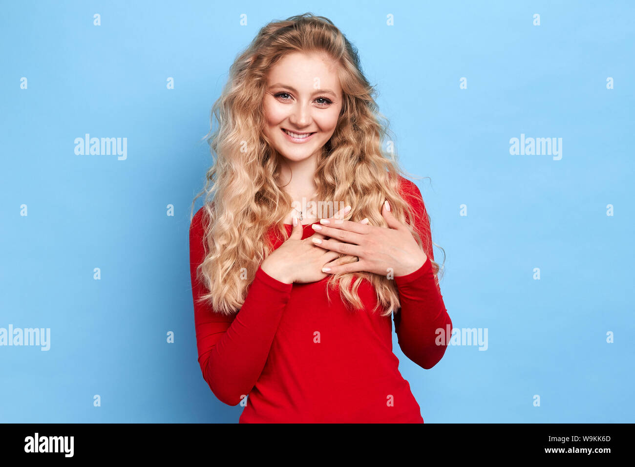 Fröhliches happy charmante romantische Mädchen hält beide Handflächen auf Herz, fühlt sich Dankbarkeit, von etwas berührt, in lässig-eleganten roten Pullover angezogen ist Stockfoto