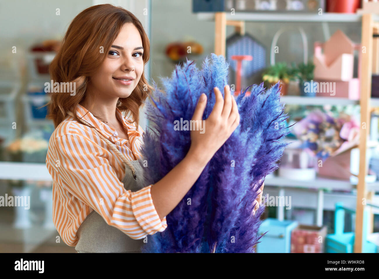 Attraktive junge Frau in stiped Shirl und Schürze umarmen Blume und posieren für die Kamera. bis Foto schliessen. Pflege, Beruf, Job Stockfoto