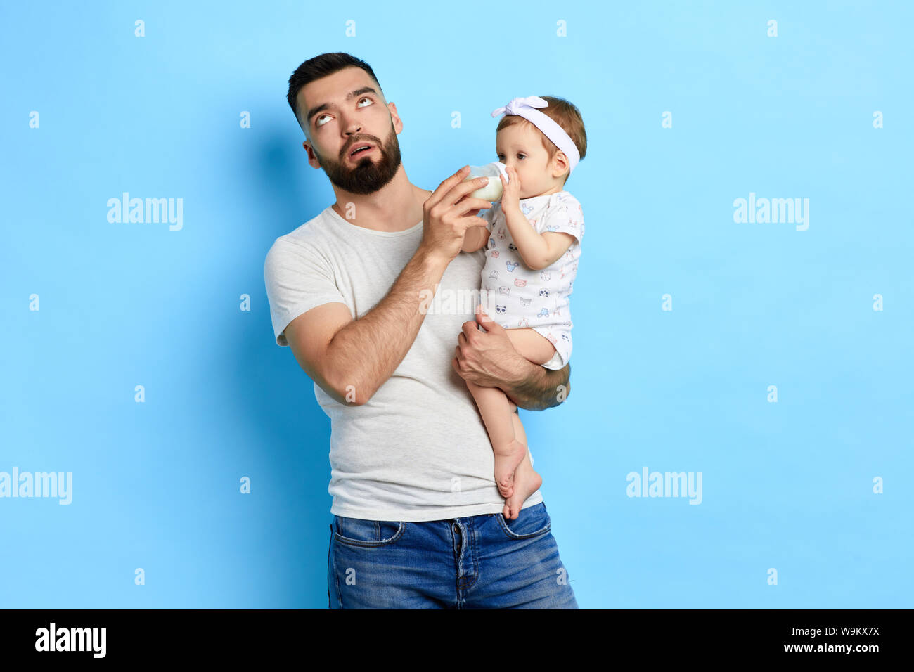 Wütend unglücklich erschöpft Vater mit einem Kind in seine Arme, ihre Milch geben, oben. Kinder kümmern. bis Foto schliessen. Isoliert blau baackground, Studio shot Stockfoto