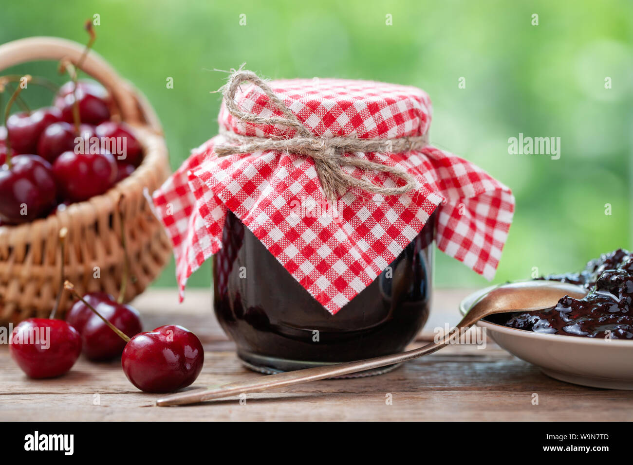 Glas Kirschen Marmelade, Korb mit Kirschen, Untertasse und Löffel auf dem Tisch im Freien. Stockfoto