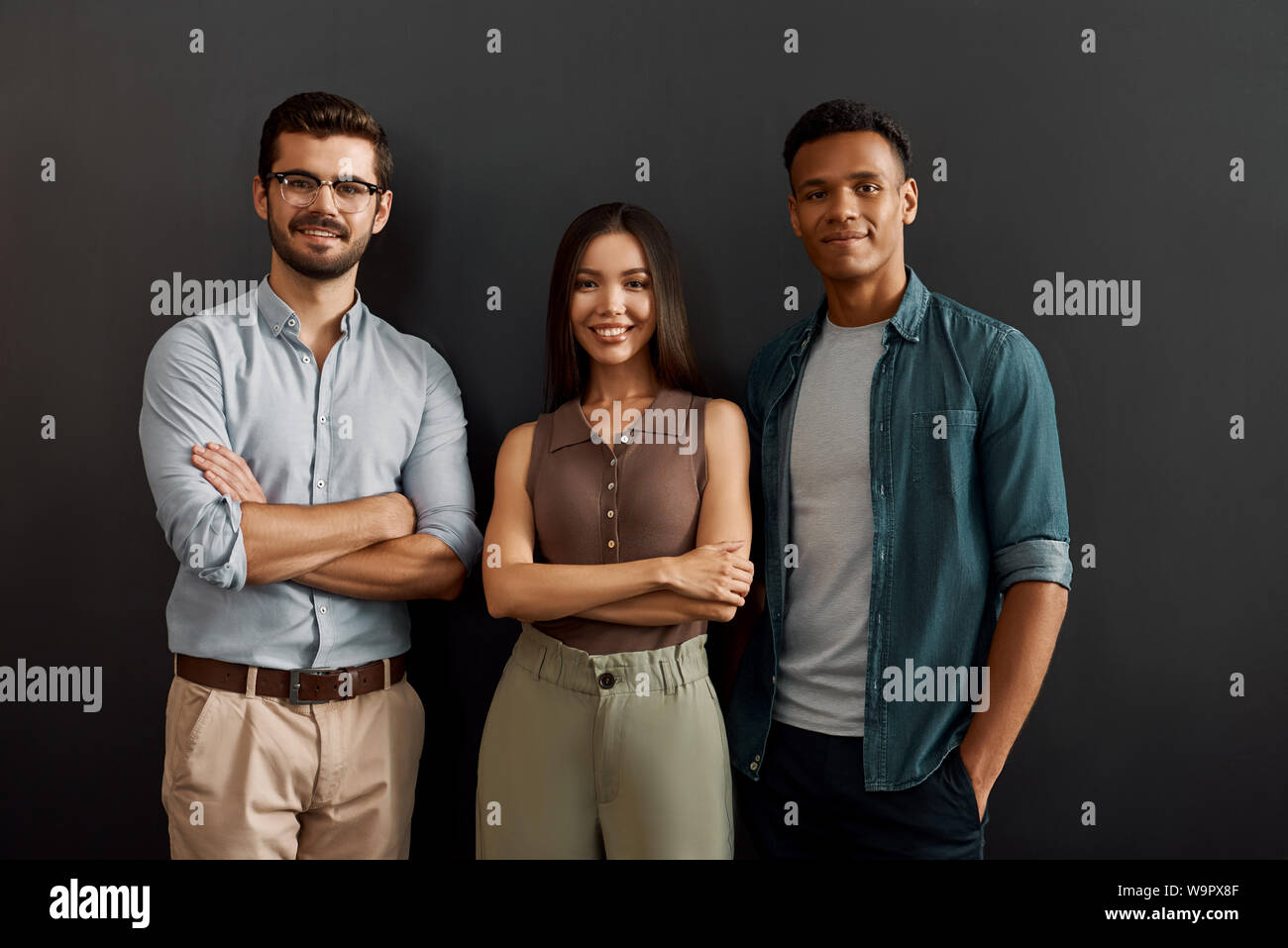 Wir sind grosse Mannschaft. Gruppe von drei multikulturellen fröhlicher junger Menschen in Freizeitkleidung auf Kamera mit Lächeln, während gegen den dunklen Hintergrund stehen. Arbeit Konzept. Zusammenarbeit. Geschäftsleute Stockfoto