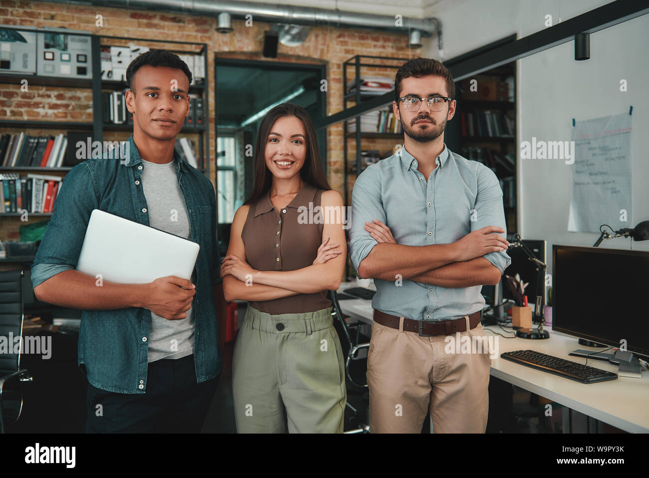 Wir sind perfekte Team. Gruppe von drei fröhliche junge Menschen in Freizeitkleidung auf Kamera mit Lächeln beim Stehen in modernen Büro. Arbeitsplatz. Business Konzept Stockfoto