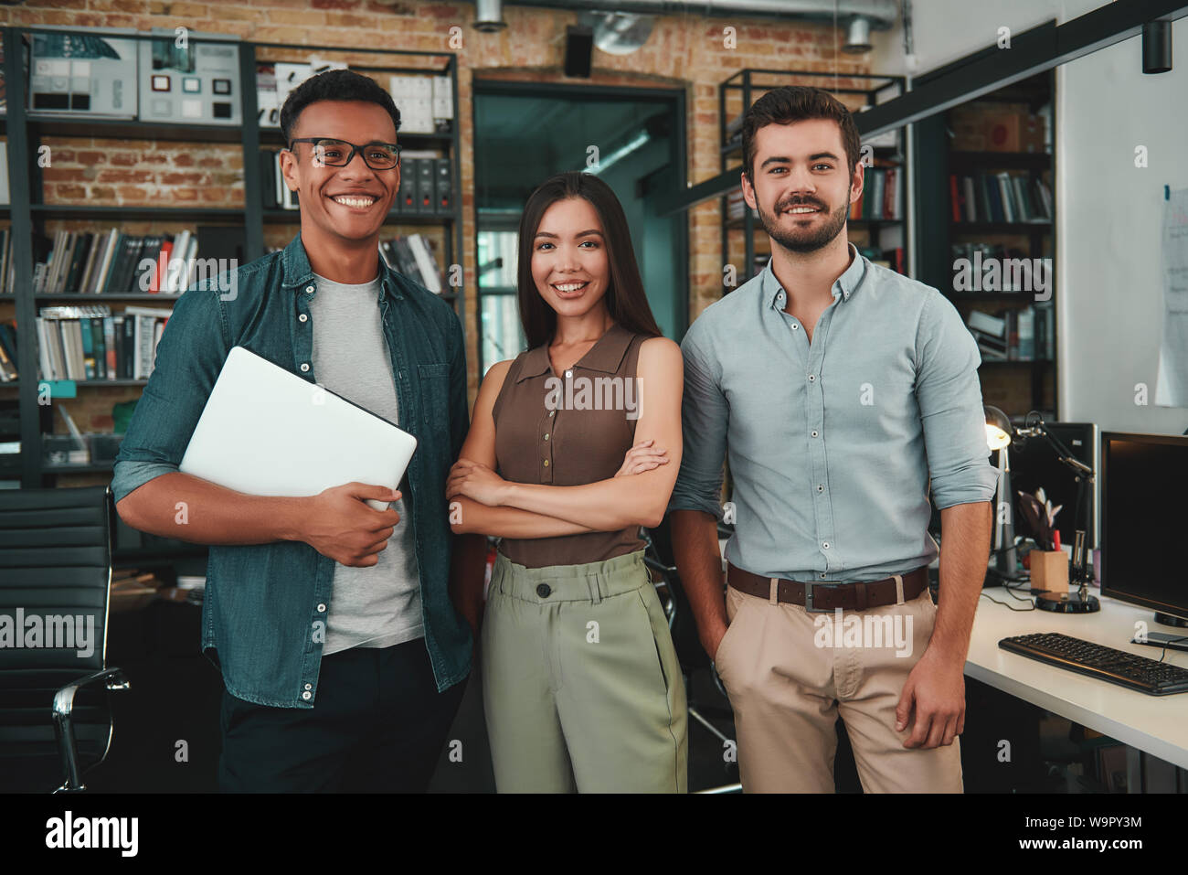 Glücklich zusammen zu arbeiten. Gruppe von drei fröhliche junge Menschen in Freizeitkleidung auf Kamera mit Lächeln beim Stehen in modernen Büro. Arbeitsplatz. Business Konzept Stockfoto