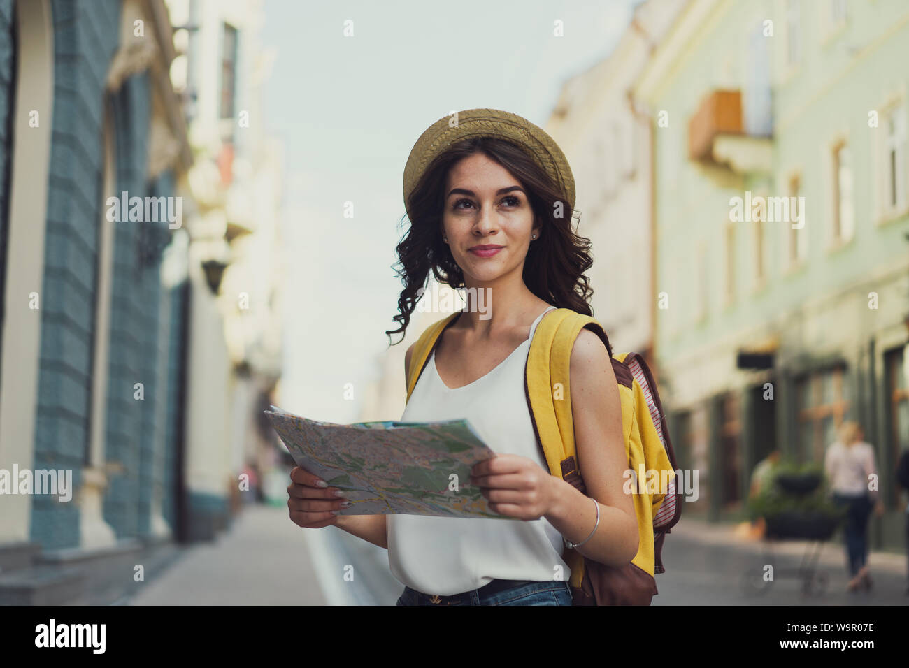 Die brünette Mädchen - Reisende mit einer Karte der Stadt und ein Rucksack in der Straße. Mit einem netten Lächeln. Und eine Architektur im Hintergrund. Stockfoto