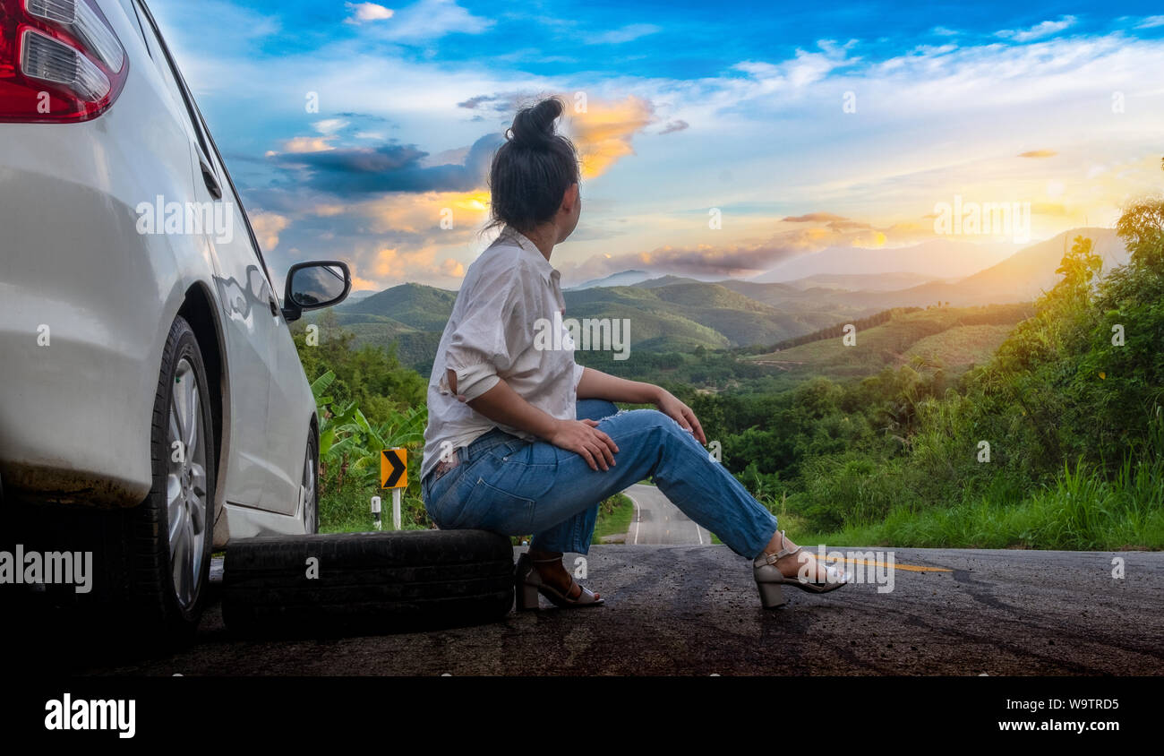 Junge hübsche Dame sitzen in der Nähe Auto für den Aufruf um Hilfe auf der öffentlichen Straße im Waldgebiet am Berg und Himmel Hintergrund Stockfoto