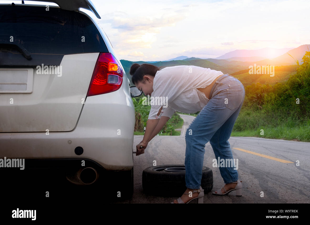 Junge attraktive Frau in formale Abnutzung Holding ein Ersatzteil Auto Reifen auf der öffentlichen Straße im Waldgebiet am Berg und Himmel Hintergrund Stockfoto