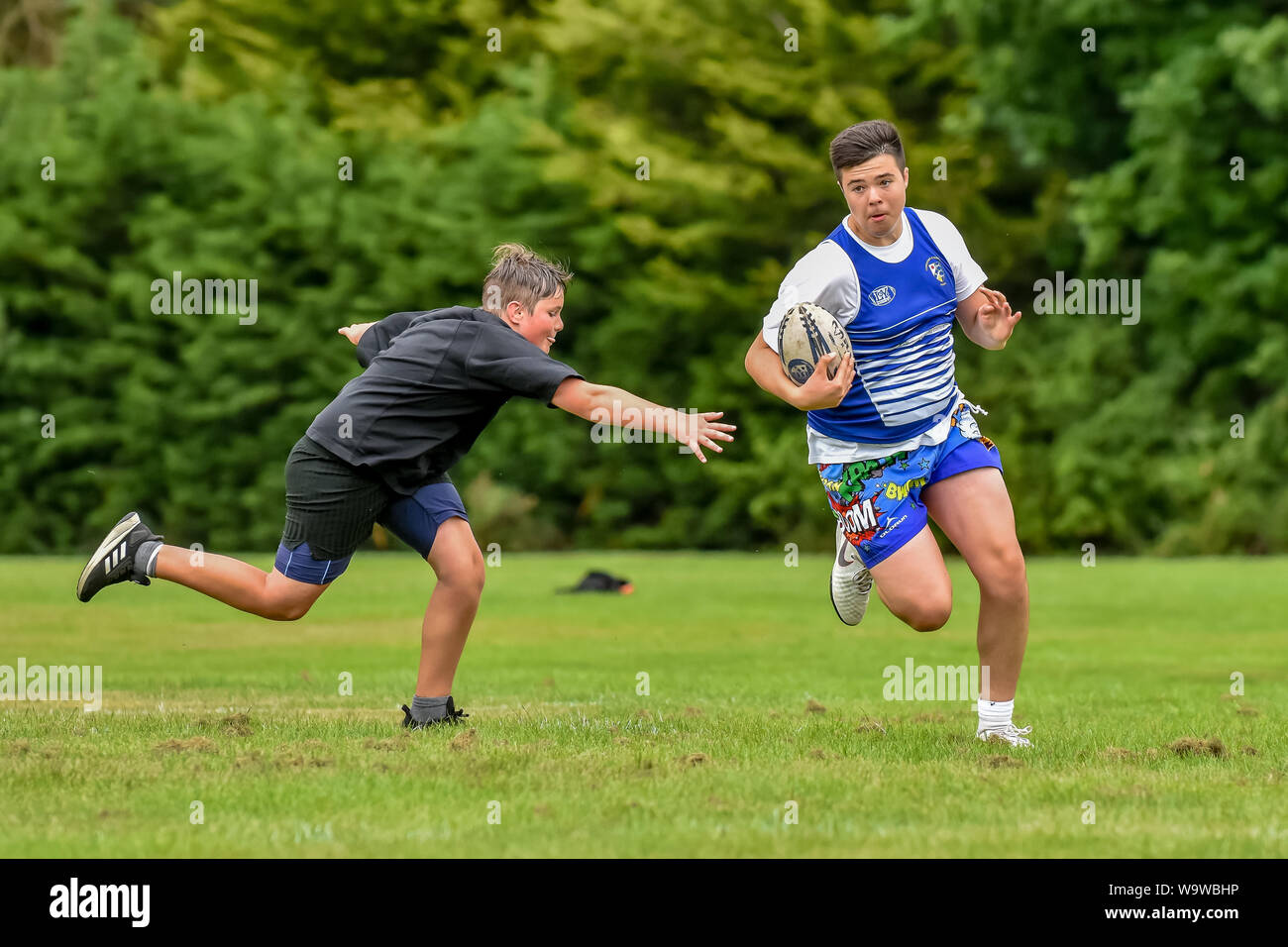 Junger Mann (Alter 15-25) sprintet mit Ball in Hand wie jüngere Spieler (Alter 14-20) erstreckt sich ein Touch zu machen am Bewunderer Touch Rugby Festival anzupacken Stockfoto