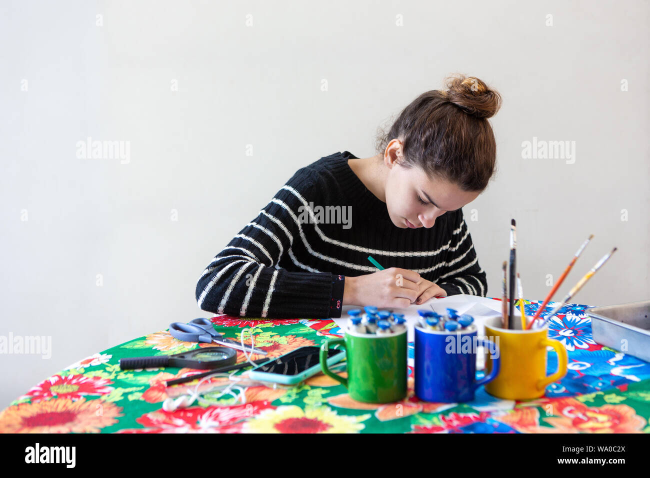 Junge talentierte Künstlerin am Tisch sitzen mit geblümten Tischdecke, Kunst mit Bleistift, Papier, Schere, Lupe, Pinsel und Farben Stockfoto