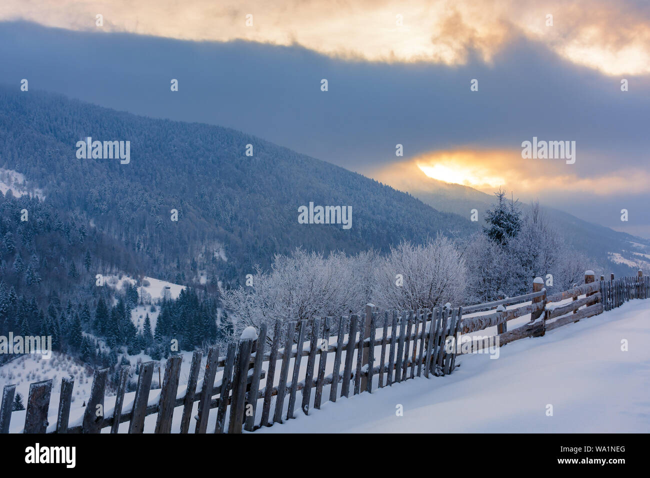 Ländliche Landschaft im Winter. schönen Sonnenaufgang mit bewölktem Himmel in den Bergen. Holz Zaun entlang der snow hill Bäume im Raureif bedeckt. Stockfoto