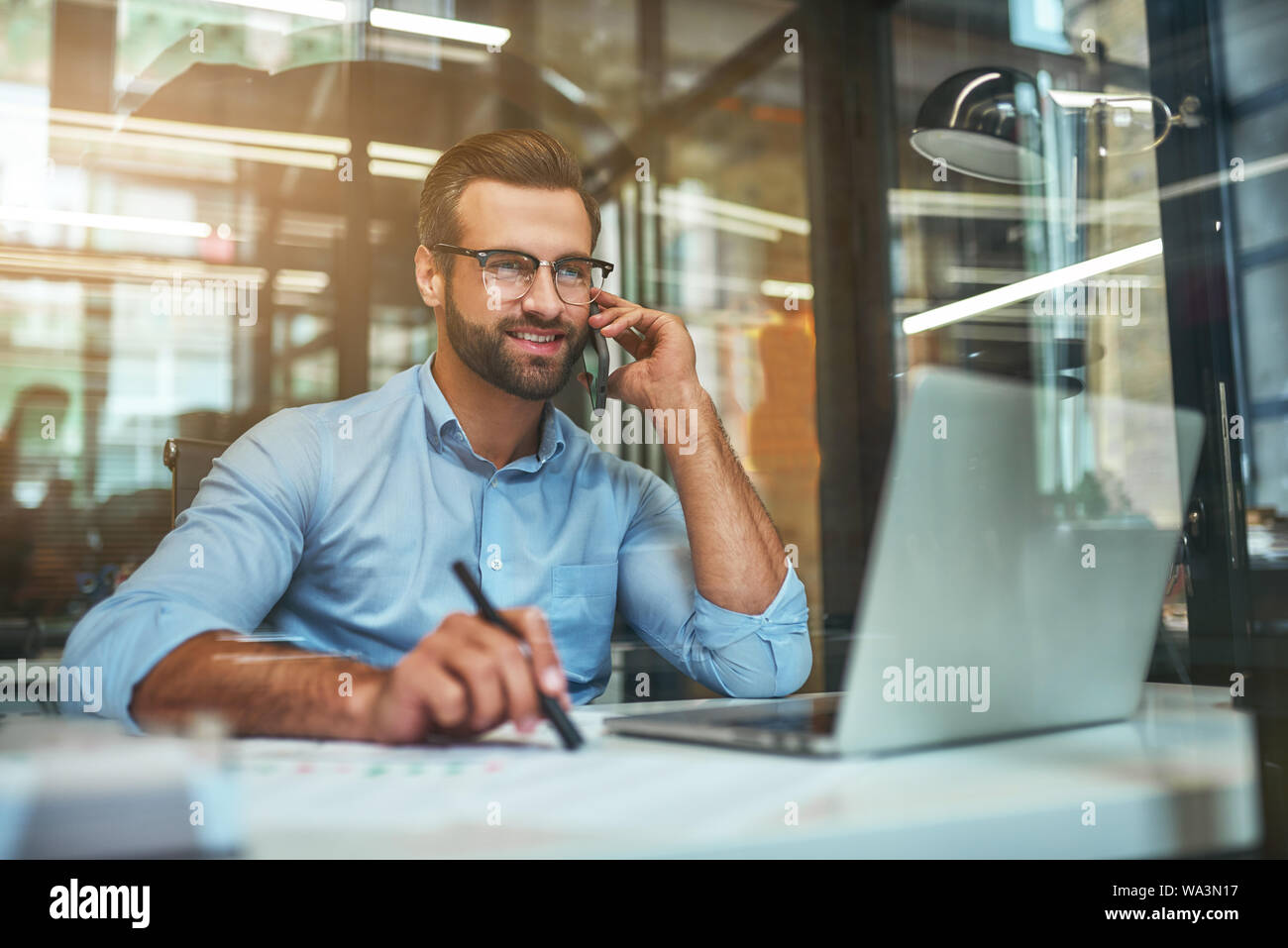 Customer Support. Porträt der jungen und freundlichen Geschäftsmann in Brillen und Gesellschaftsmode, am Telefon zu sprechen und lächelnd, während im Büro zu sitzen. Arbeitsplatz. Business Konzept Stockfoto