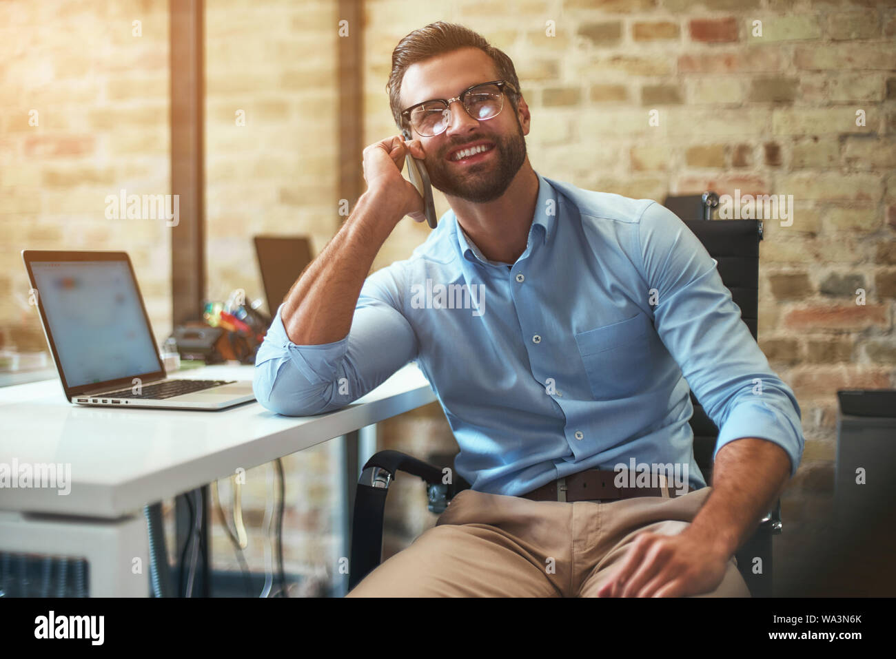 Tolle Neuigkeiten Portrait von junge Bartgeier Geschäftsmann in Brillen und Gesellschaftsmode, am Telefon zu sprechen und lächelnd, während in der komfortablen Büro zu sitzen. Geschäftskonzept. Kommunikation Stockfoto