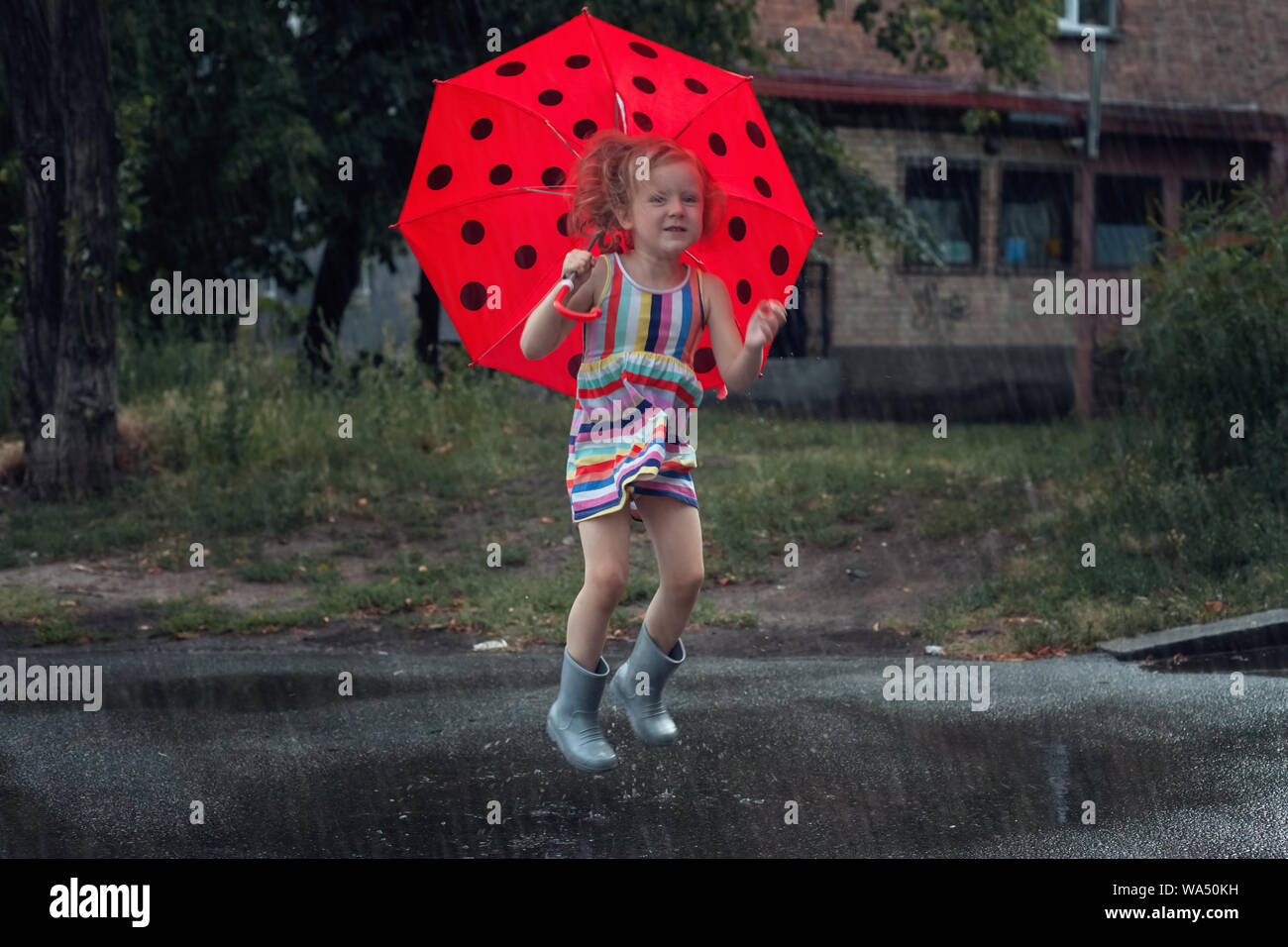 Kleines Mädchen mit Schirm springen im Regen durch die Pfützen. Stockfoto