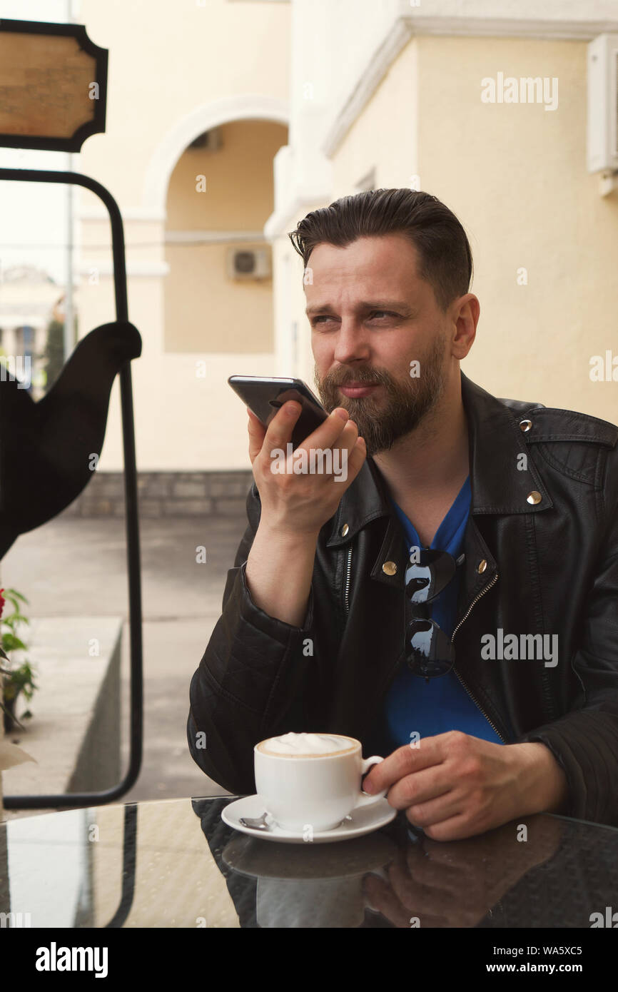 Stilvolle hipster Freiberufler Kaffee trinken auf der Terrasse eines Restaurants. Ein Mann ist Aufnahme einer Sprachnachricht. Hübscher Kerl mit einem modischen ha Stockfoto