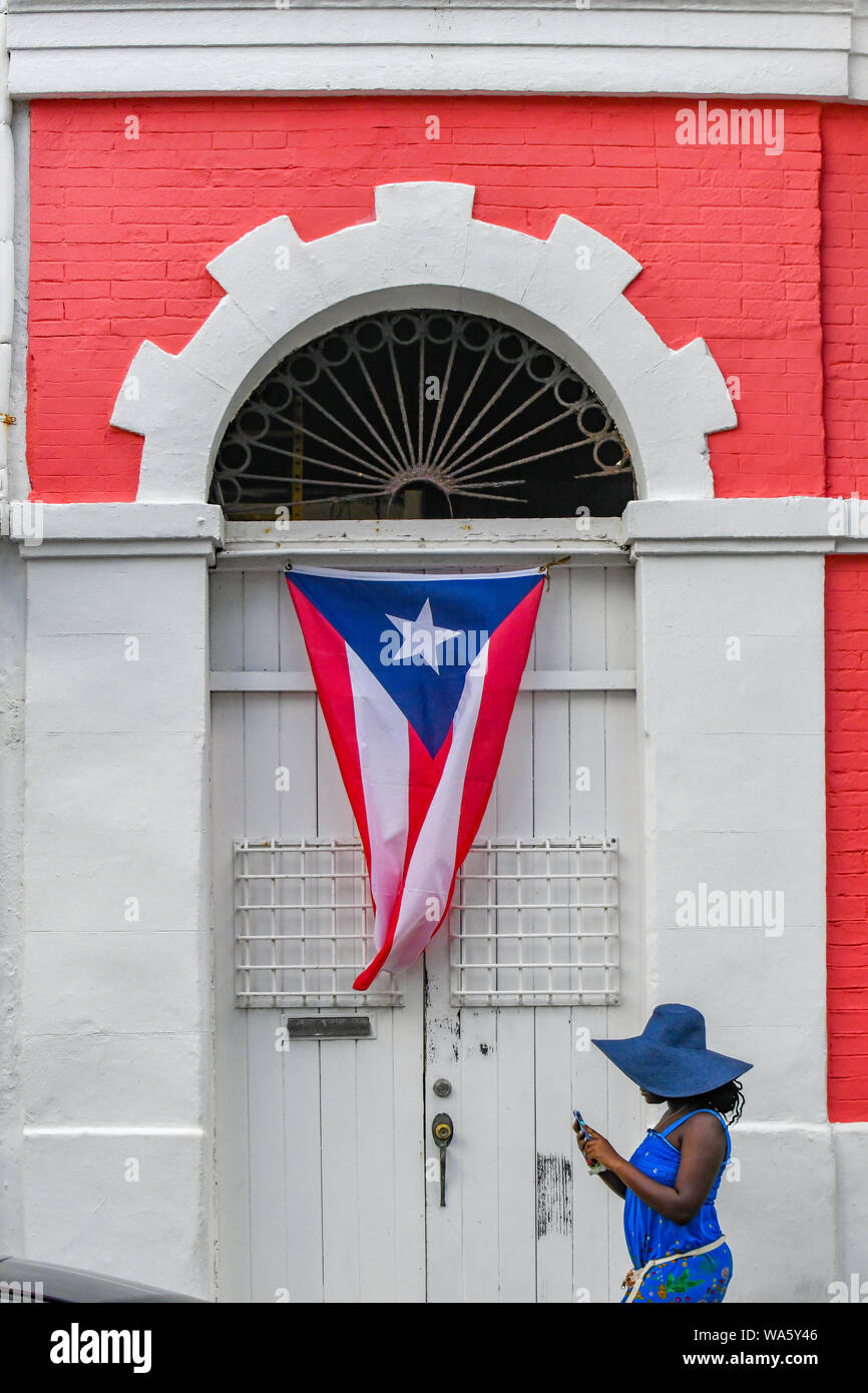 Puerto Rico Fahne flattert - eine Frau in einem blauen Kleid mit einem Handy Wanderungen von einem bunten Gebäude in der Innenstadt von Old San Juan Puerto Rico Flagge haengt Stockfoto