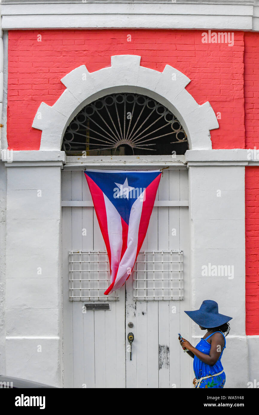 Puerto Rico Fahne flattert - eine Frau in einem blauen Kleid mit einem Handy Wanderungen von einem bunten Gebäude in der Innenstadt von Old San Juan Puerto Rico Flagge haengt Stockfoto