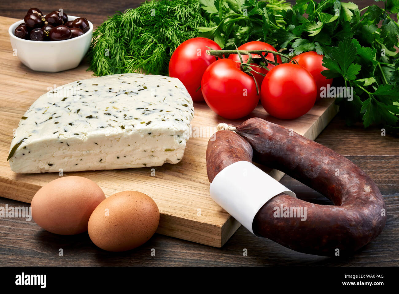 Türkisch Van Milch Käse mit Kräutern auf Holztisch mit Holz Schneidebrett mit fermentierten Würstchen, Eier, Olivenöl, Petersilie und Tomaten. Stockfoto