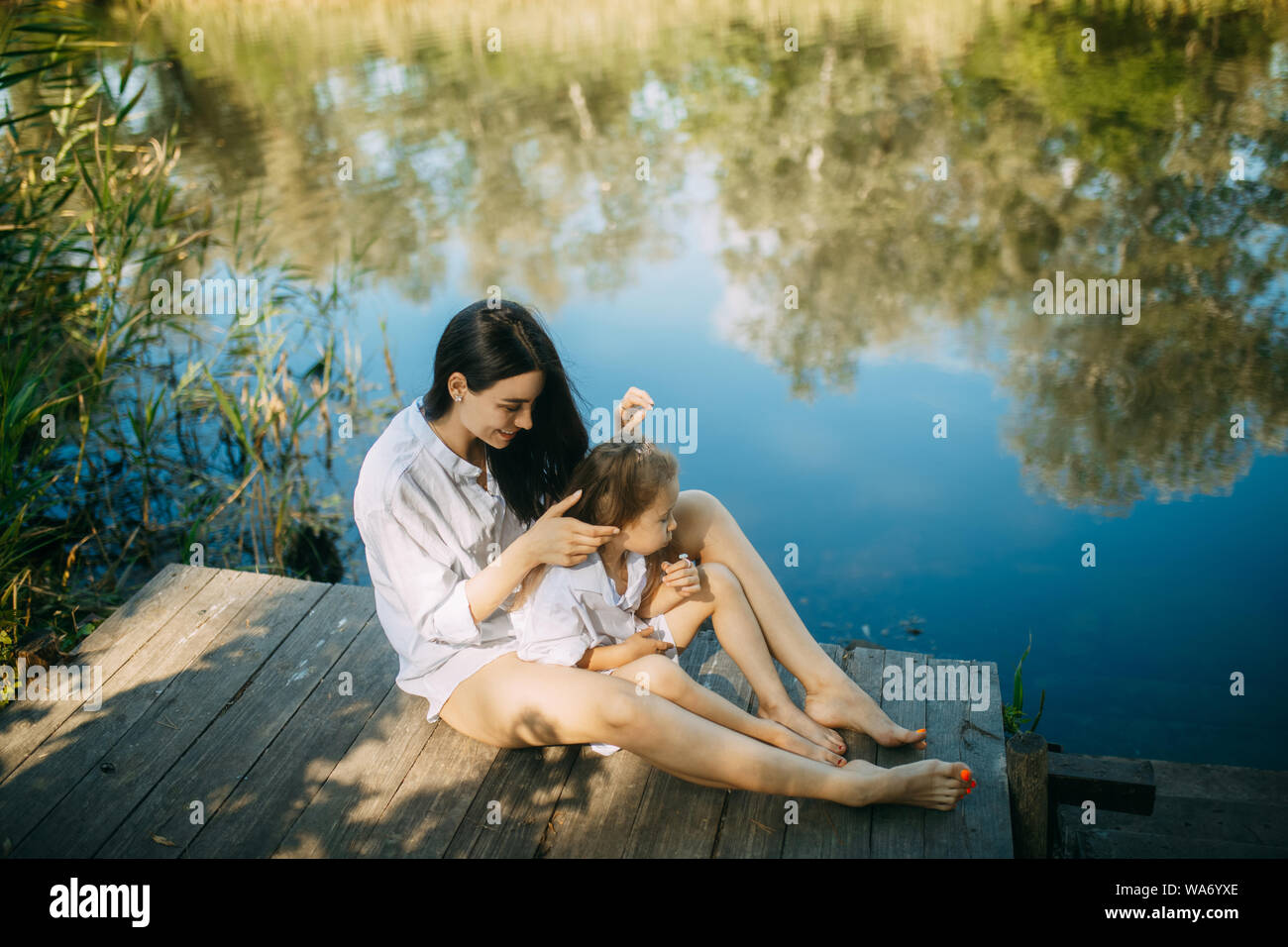 Mutter und Tochter sitzen auf einem hölzernen Brücke über einen Fluss mit Reflexion der Bäume und der Himmel im Wasser. Stockfoto