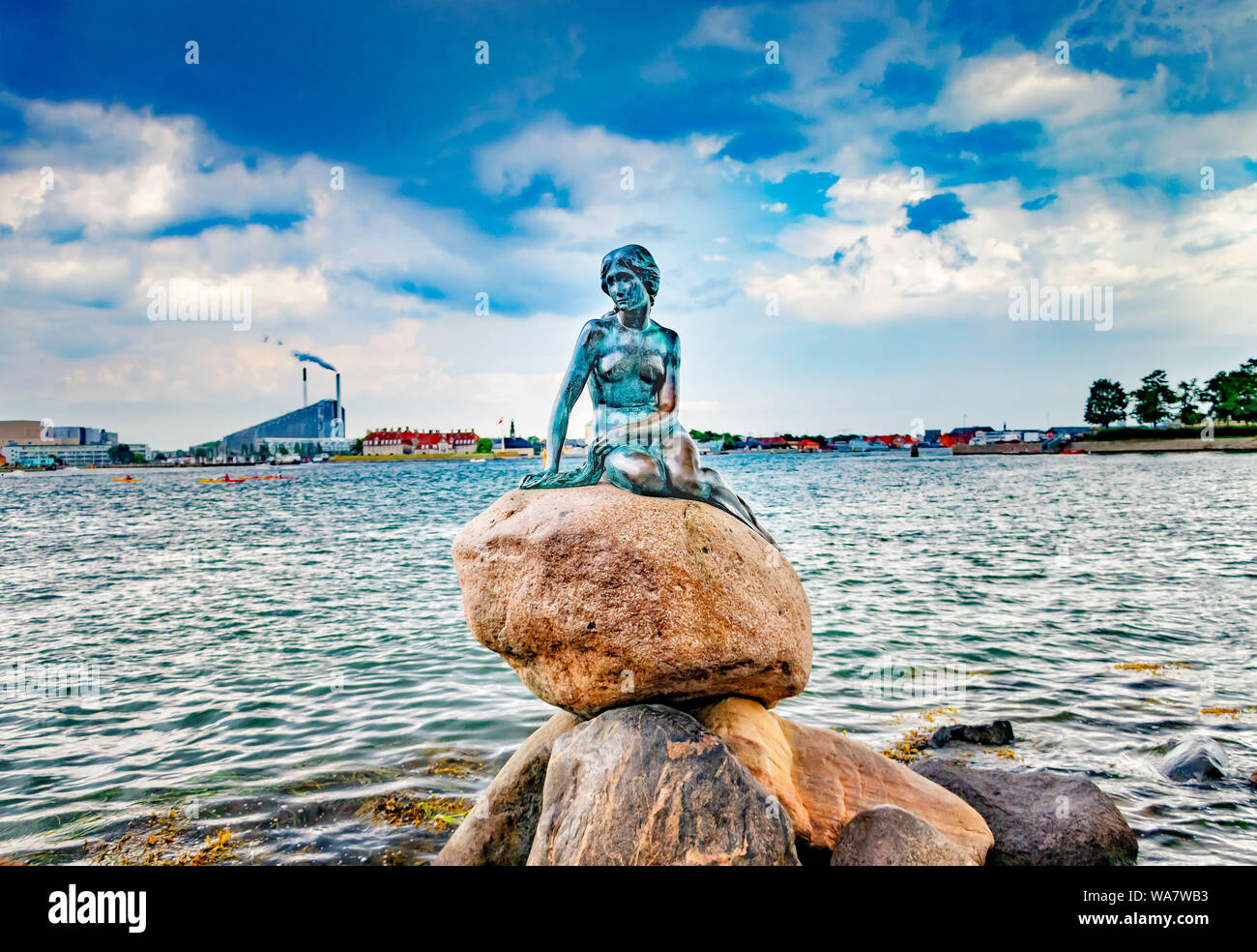 Statue der kleinen Meerjungfrau sitzend auf Stein. Dahinter ist das Meer und die Aussicht auf den Hafen von Kopenhagen. Stockfoto