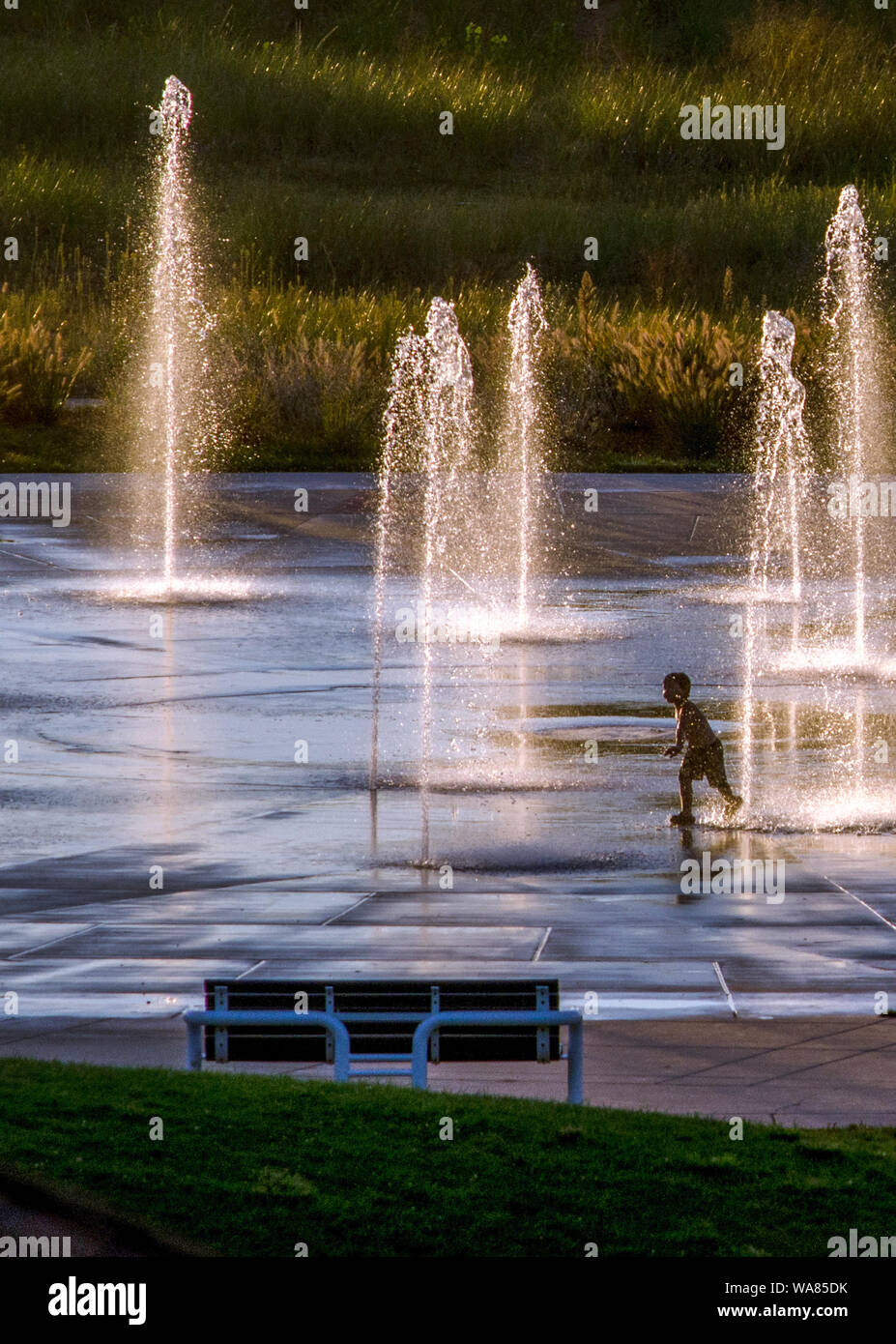 Kleiner Junge läuft glücklich in einem Urban Splash Pad bei Dämmerung Stockfoto