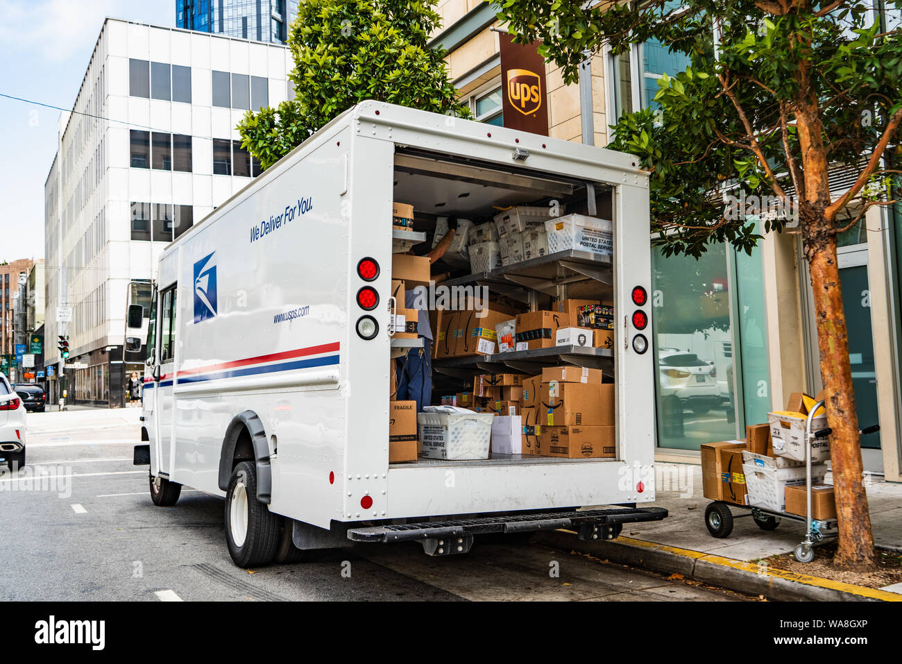 August 10, 2019 San Francisco/CA/USA - USPS delivery van vor einem UPS gestoppt, entladen Amazon Pakete Stockfoto