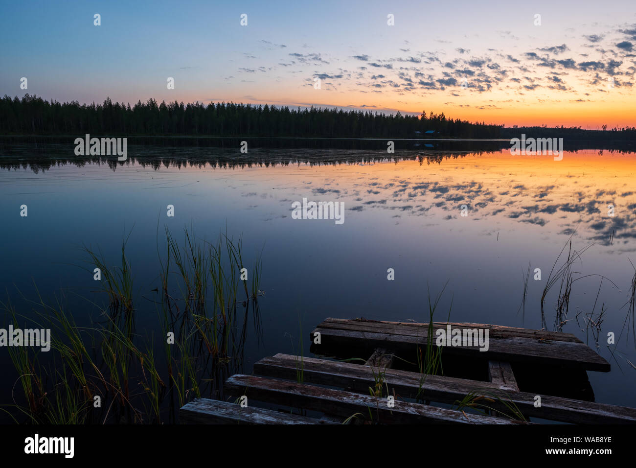 Sonnenuntergang und Einsamkeit an einem finnischen See mit schönen Reflexionen von Wolken und Dunkel borealen Wald und alte Pier im Vordergrund, Finlan Stockfoto