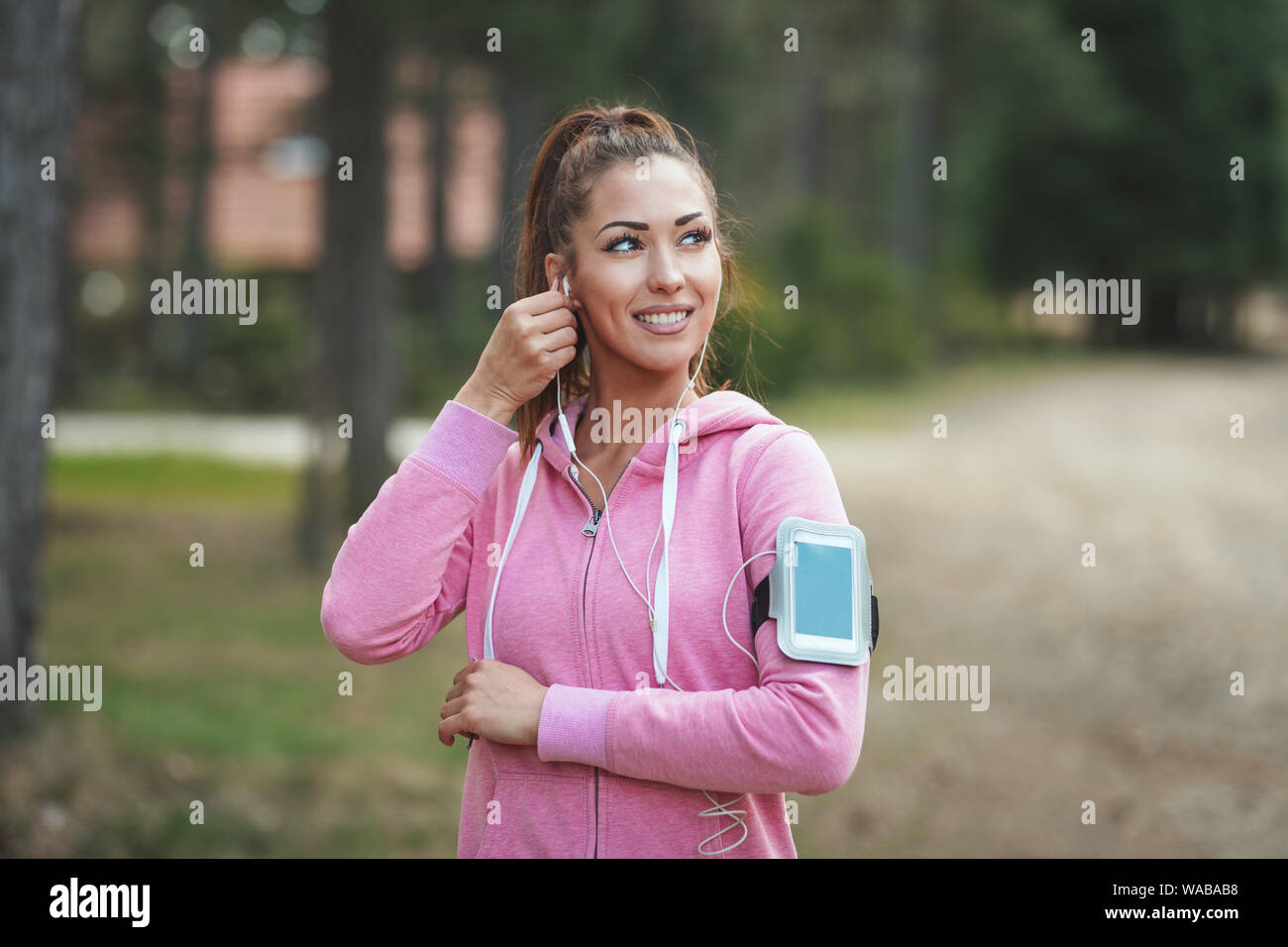 Glückliche Frau, Sportkleidung tragen, mit Kopfhörern in den Ohren, ist das Hören von Musik in einer Pause von Übungen an einem sonnigen Wald, genießen Landschaft zwischen Th Stockfoto