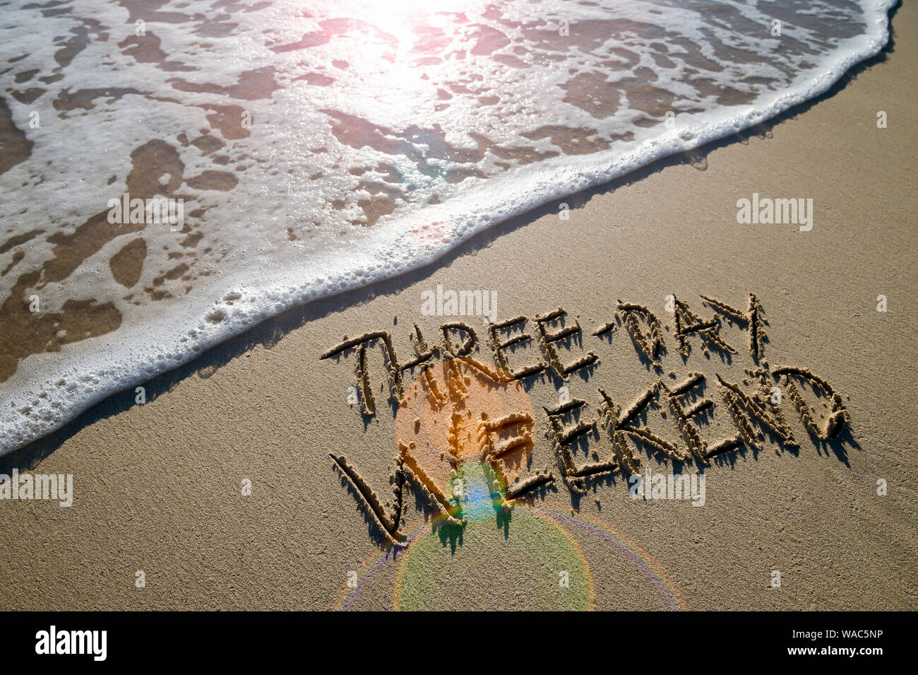 Three-Day Wochenende Nachricht handschriftlich auf glatten Sandstrand mit Lens Flare über einen ankommenden Welle Stockfoto